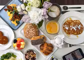 Et morgenmadspålæg med røræg, sprød bacon, diverse kager, pandekager med pulveriseret sukker, frugt, yoghurt med nødder, et kogt æg og en frugtdrik. En buket blomster, en cappuccino og en smartphone er også synlige på bordet.