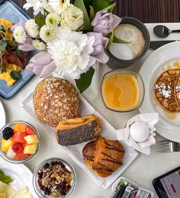 Et morgenmadspålæg med røræg, sprød bacon, diverse kager, pandekager med pulveriseret sukker, frugt, yoghurt med nødder, et kogt æg og en frugtdrik. En buket blomster, en cappuccino og en smartphone er også synlige på bordet.
