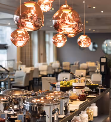 Et moderne spiseområde med en buffetopstilling med gnidningsfade i rustfrit stål, forskellige madvarer og frugt. Kobber pendler hænger ovenover, og baggrunden viser pænt arrangerede borde og stole i et godt oplyst rum.