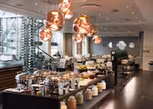 En moderne restaurantbuffet med et poleret trægulv. Der er skinnende lysekroner og krukker med korn og krydderier på en central ø. Buffeten har gnavende retter med forskellige fødevarer. Spiseområdet er sat med hvide stole og borde.