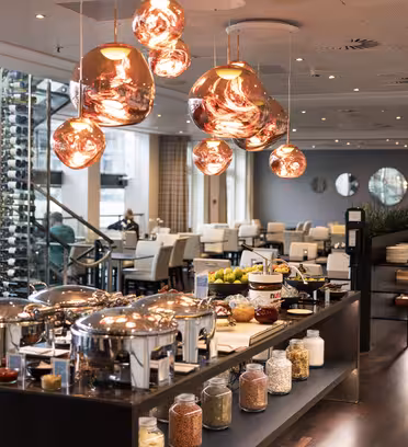 En moderne restaurantbuffet med et poleret trægulv. Der er skinnende lysekroner og krukker med korn og krydderier på en central ø. Buffeten har gnavende retter med forskellige fødevarer. Spiseområdet er sat med hvide stole og borde.