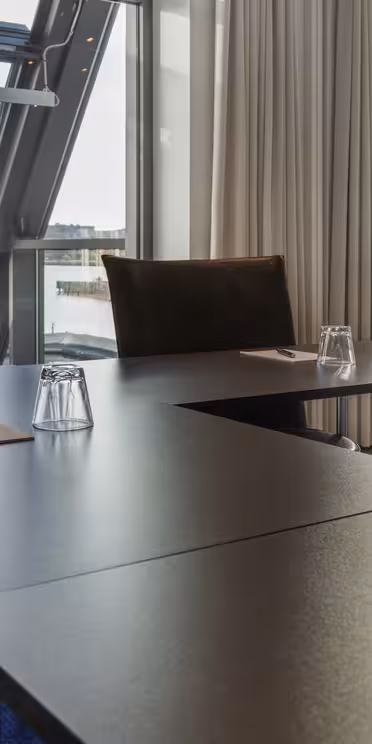 Et moderne mødelokale med et U-formet bordarrangement. Stole er sat omkring borde med mørkt tema, og hver indstilling inkluderer en notesblok og glas. Store vinduer giver udsigt til havnefronten og bybilledet udenfor, hvilket giver naturligt lys mulighed for at fylde rummet.