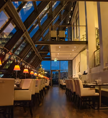 Elegant restaurant med glasloft og store vinduer, der viser et bybillede i skumringen. Interiøret byder på beige stole, trægulve, hængende dekorationer og varm belysning, der skaber en hyggelig atmosfære.