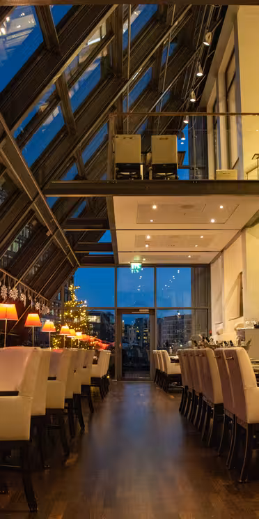 Elegant restaurant med glasloft og store vinduer, der viser et bybillede i skumringen. Interiøret byder på beige stole, trægulve, hængende dekorationer og varm belysning, der skaber en hyggelig atmosfære.