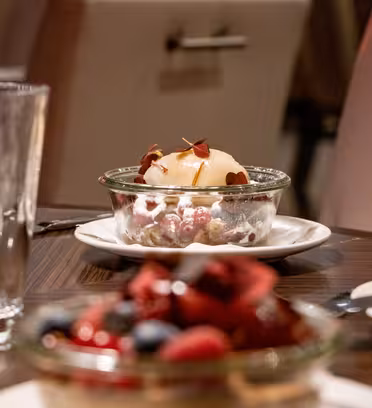 Et dessertbord med to glasskåle med desserter. Det nærmeste fad er fyldt med blandede bær, og jo længere man har en kugle is pyntet med blade. Et glas vand og bestik står på træbordet.