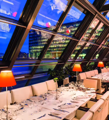 En restaurant med en skrå glasvæg med udsigt over bybilledet ved vandet om natten. Interiøret byder på hvide polstrede stole, pænt dækkede borde med glas og sølvtøj og røde lamper, der skaber en varm og elegant atmosfære.