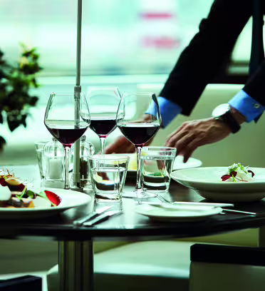 En person i jakkesæt dækker et bord med tallerkener med gourmetmad, vinglas fyldt med rødvin og vandglas. Bordet er elegant arrangeret i moderne restaurant-miljø med en plante i baggrunden.