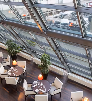 Moderne restaurantinteriør med glasvægge, der giver udsigt til en marina. Hvide polstrede stole omkranser runde træborde med orange lamper. Store grønne planter tilføjer indretningen.