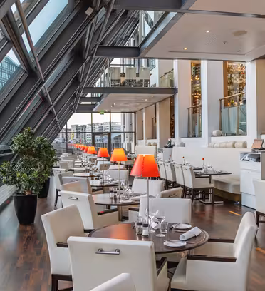 Moderne restaurantinteriør med hvide polstrede stole og mørke træborde sat med glasvarer og servietter. Store skrå vinduer langs den ene side og giver udsigt til bygninger udenfor. Røde bordlamper tilføjer et varmt skær i hele rummet.