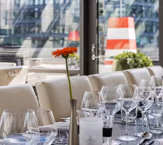 Elegant spisestue med hvide borddækninger og glasvarer på et langt, mørkt bord. To orange gerbera-margueritter i vaser tilføjer et glimt af farve. Store vinduer afslører en urban baggrund med en rød og hvid stribet struktur synlig udenfor.