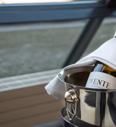 En flaske vin hviler i en metalisspand, delvist pakket ind med en hvid serviet. Flasken har en synlig etiket med tekst. Baggrunden viser et sløret, moderne indre rum med glas og strukturelle elementer.