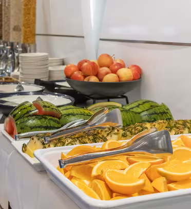 Et buffetbord med skiver af appelsiner, vandmelon og ananas. Skåle og serveringstang sættes på en hvid dug. En skål æbler og dispensere med korn er i baggrunden. Plader er stablet i nærheden.