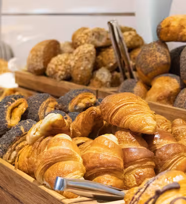 En række forskellige kager og brød er stablet i træbakker. Sortimentet omfatter croissanter, rundstykker toppet med valmuefrø og andet bagværk, pænt arrangeret med tang til servering.